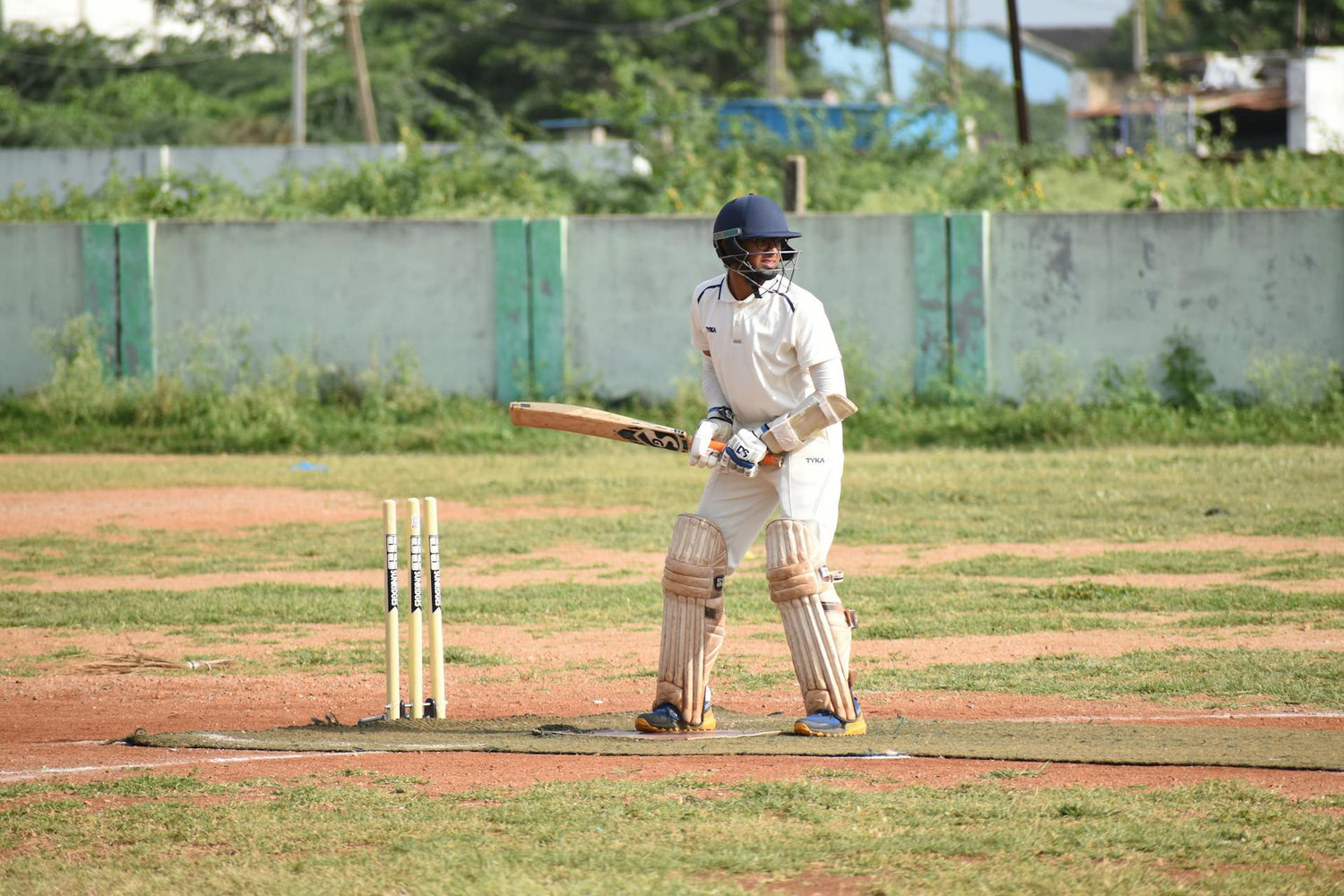 Cricketer wearing protective gear and a uniform by Trustline Enterprises, standing ready to bat on a cricket field.