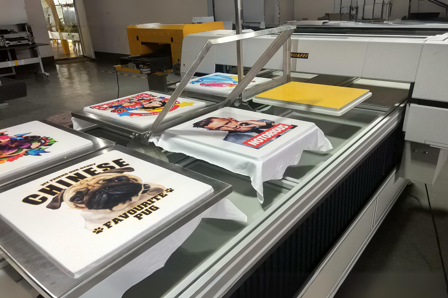 Printing machine in a workshop producing custom t-shirts with various colorful designs by Trustline Enterprises.