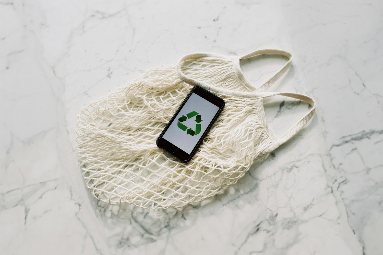 An eco-friendly reusable net bag with a smartphone displaying a recycling symbol, placed on a marble surface.