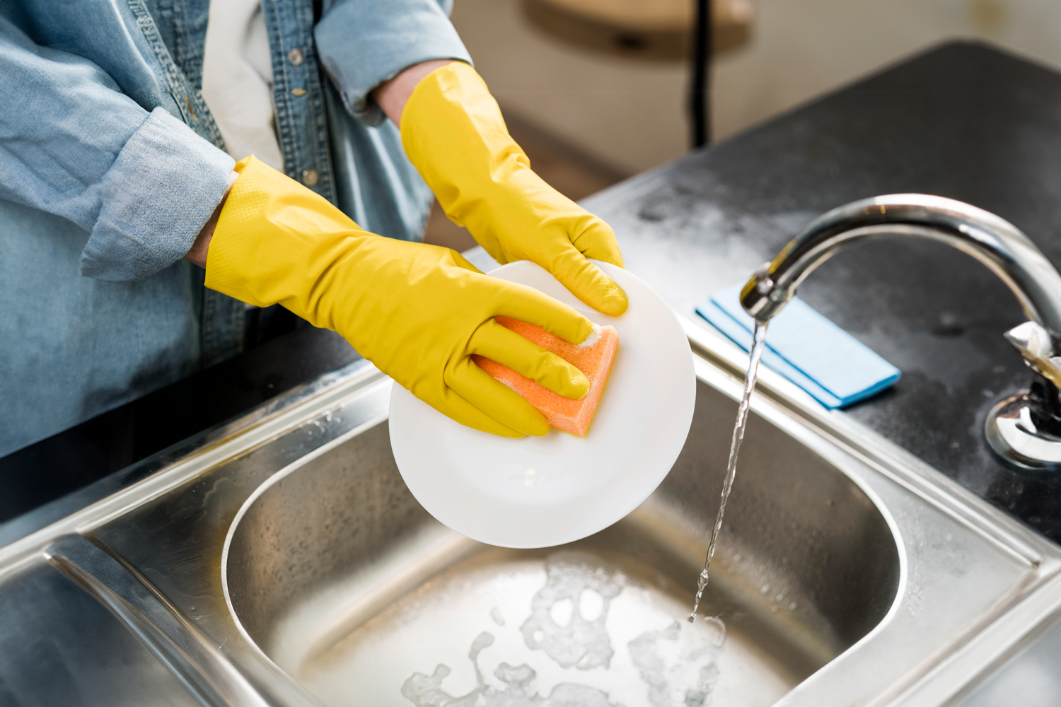 Person wearing yellow gloves washing dishes with a sponge at a kitchen sink, representing Trustline Enterprises' high-quality cleaning supplies for foodservice and hospitality industries.