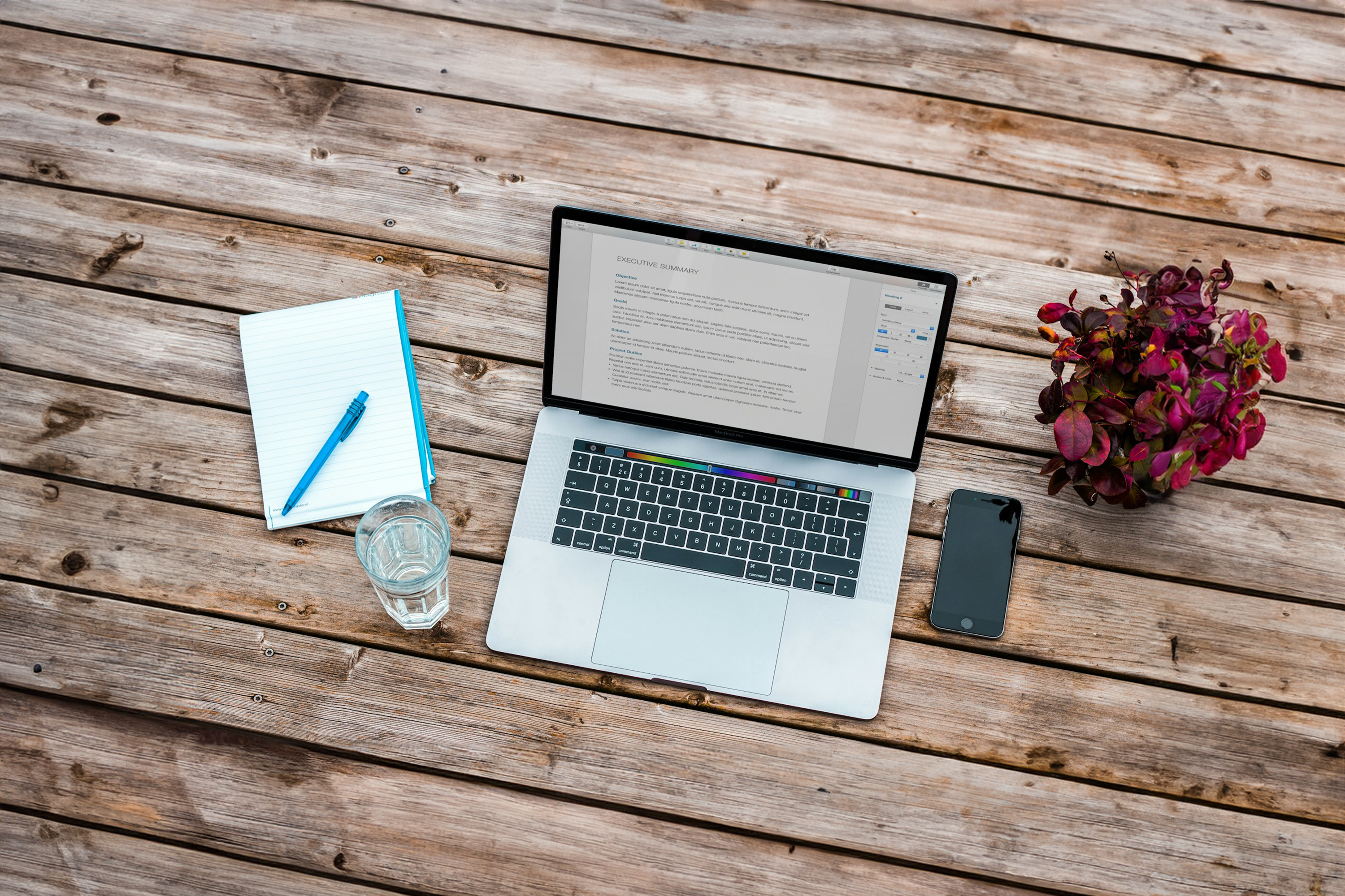 A wooden desk with a laptop displaying a document, a notepad with a pen, a glass of water, a smartphone, and a small potted plant with red leaves.