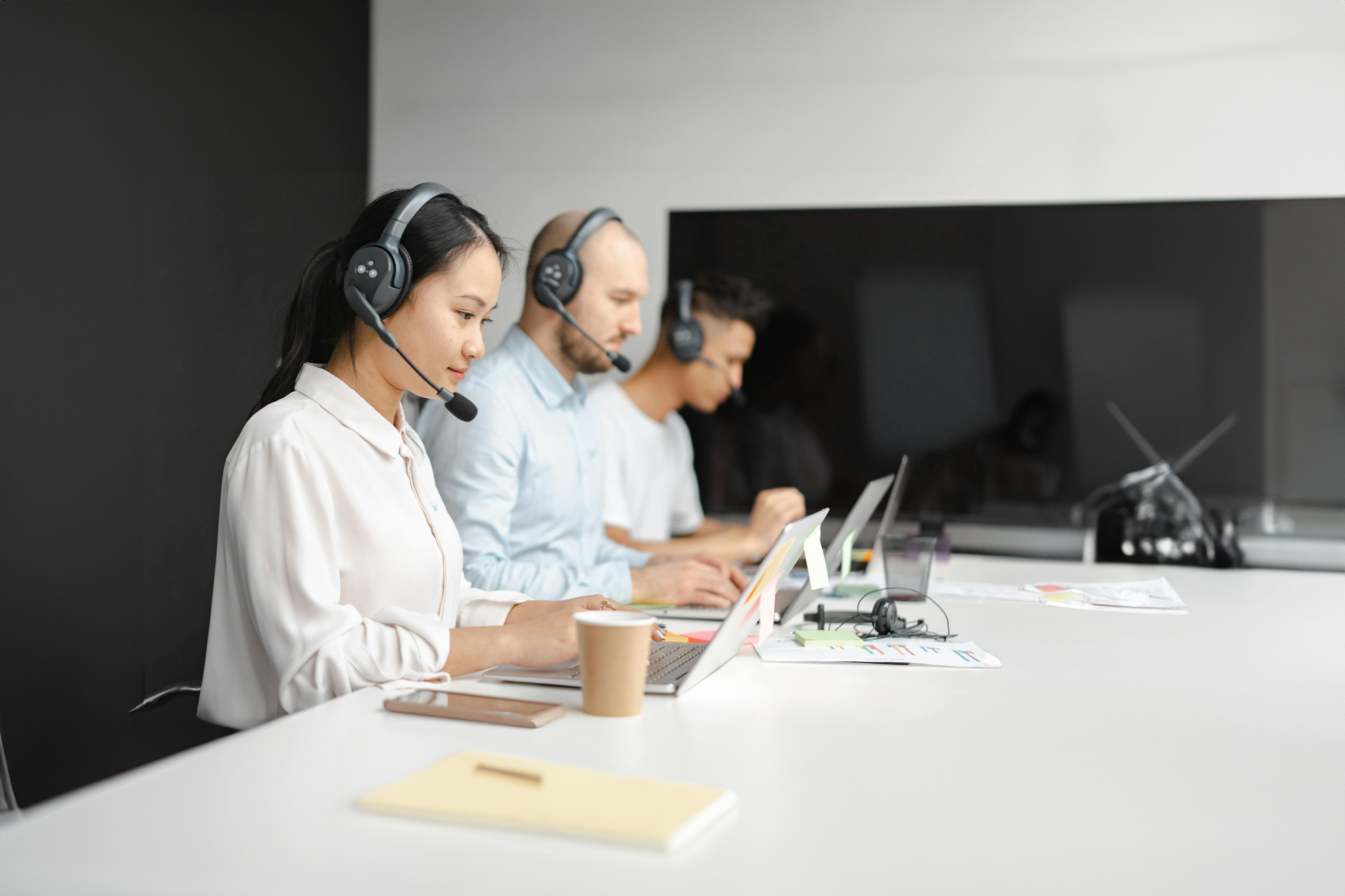 Customer service representatives wearing headsets, sitting in a row at a desk with laptops, working in a modern office environment.