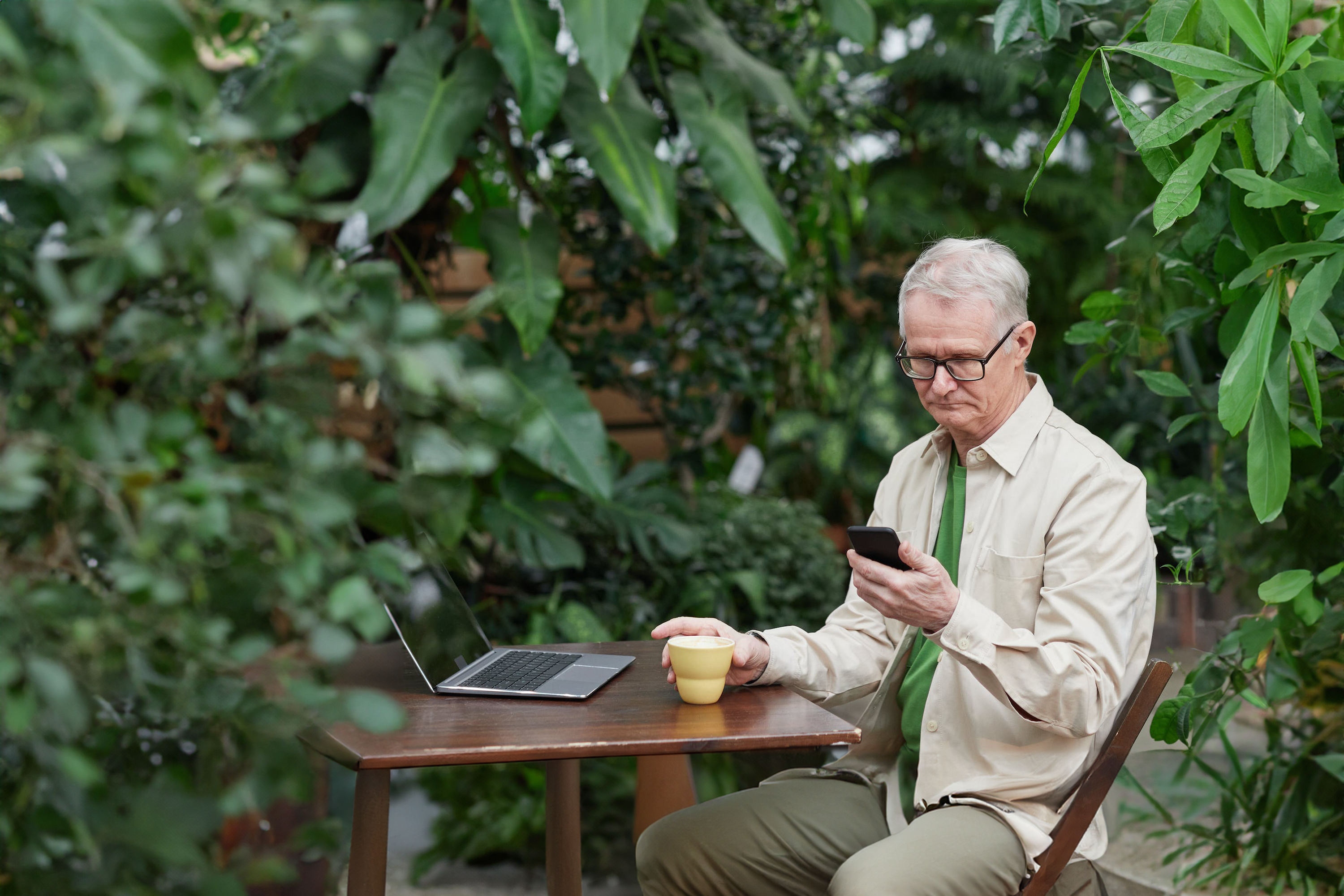 Elderly man sitting at an outdoor table surrounded by greenery, using a smartphone and holding a coffee mug, with a laptop open in front of him.