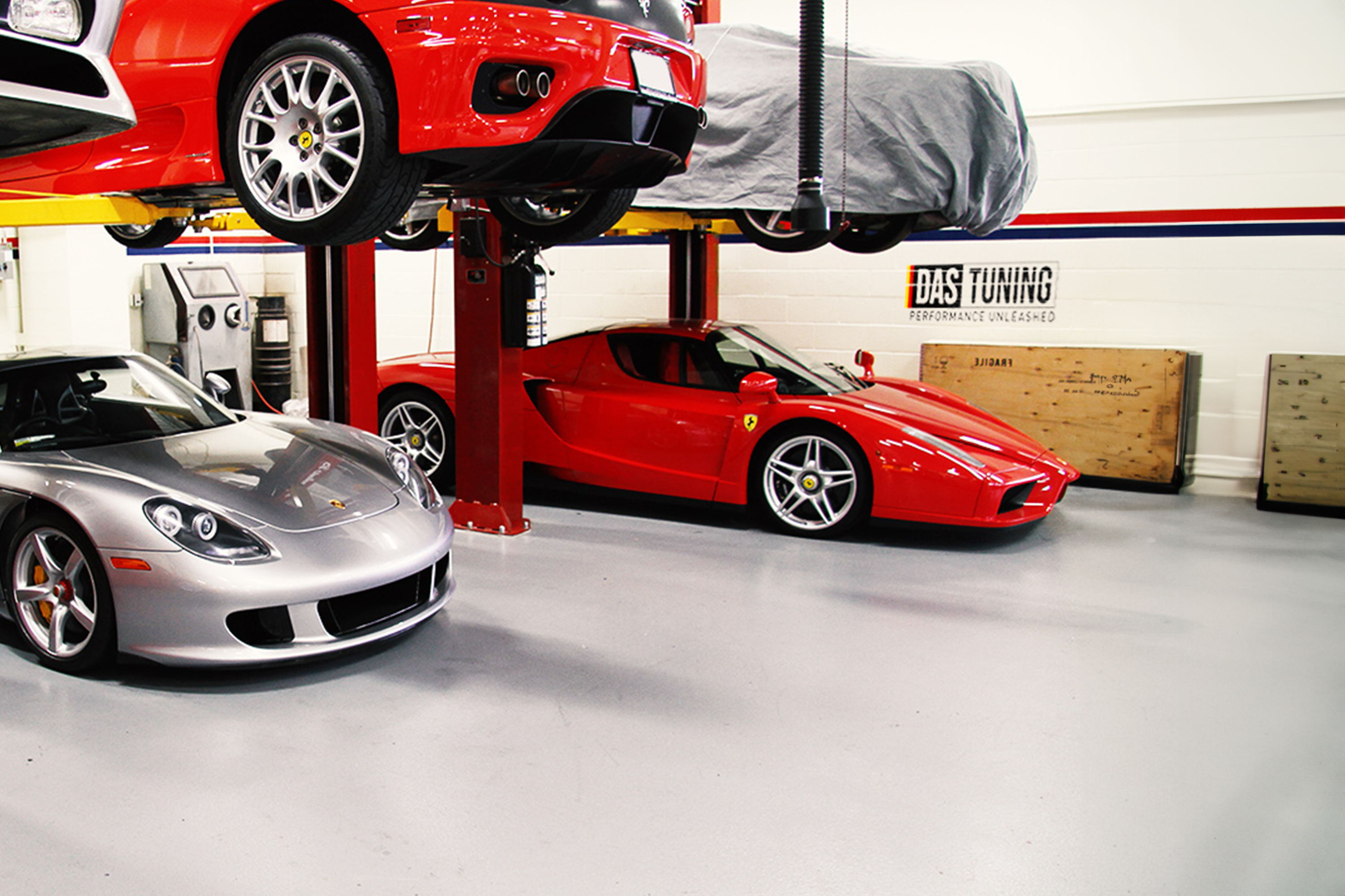 Interior of a high-end car tuning and performance shop featuring luxury sports cars, including a red Ferrari and a silver Porsche, elevated on hydraulic lifts. The shop is well-organized and clean, showcasing the professionalism and expertise of DAS Tuning.