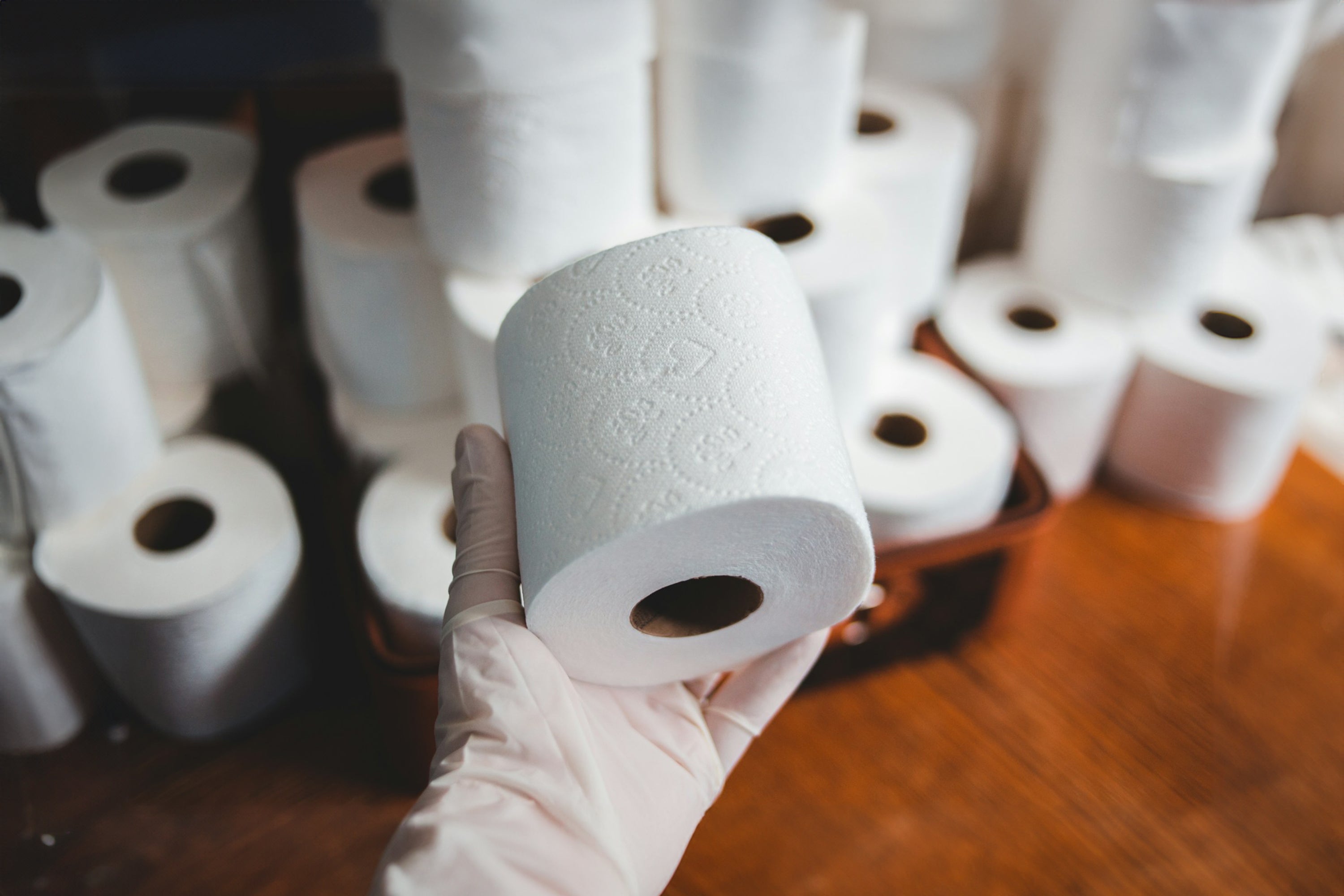 Close-up of a person wearing a glove, holding a roll of embossed toilet paper. In the background, there are multiple rolls of toilet paper stacked and scattered on a wooden surface, suggesting bulk storage or stockpiling.