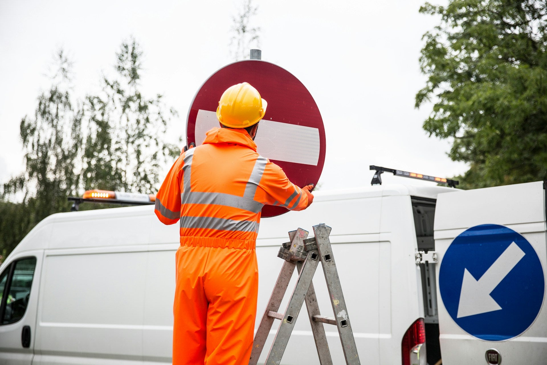 Construction worker in high-visibility orange gear and a hard hat placing a no-entry road sign next to a white van, with a ladder and trees in the background.