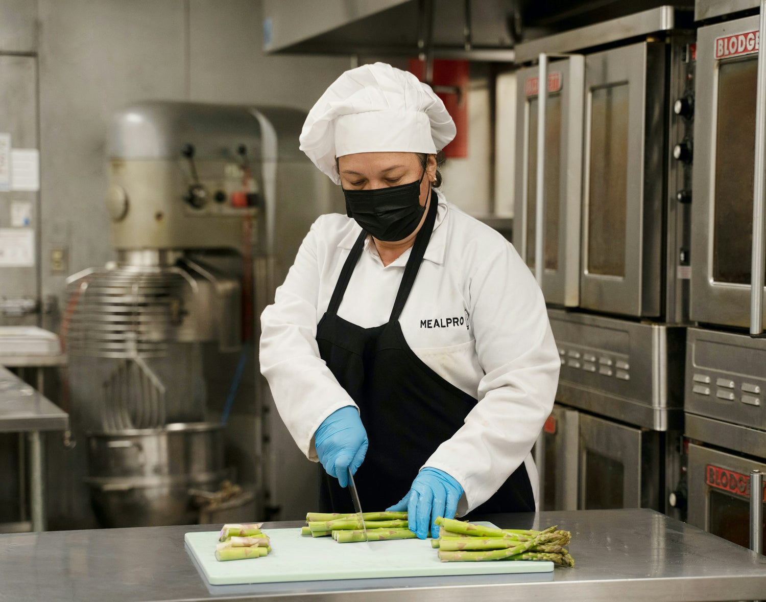 Chef wearing a white uniform, black apron, and face mask, cutting asparagus on a green cutting board in an industrial kitchen setting, with stainless steel appliances and equipment in the background.