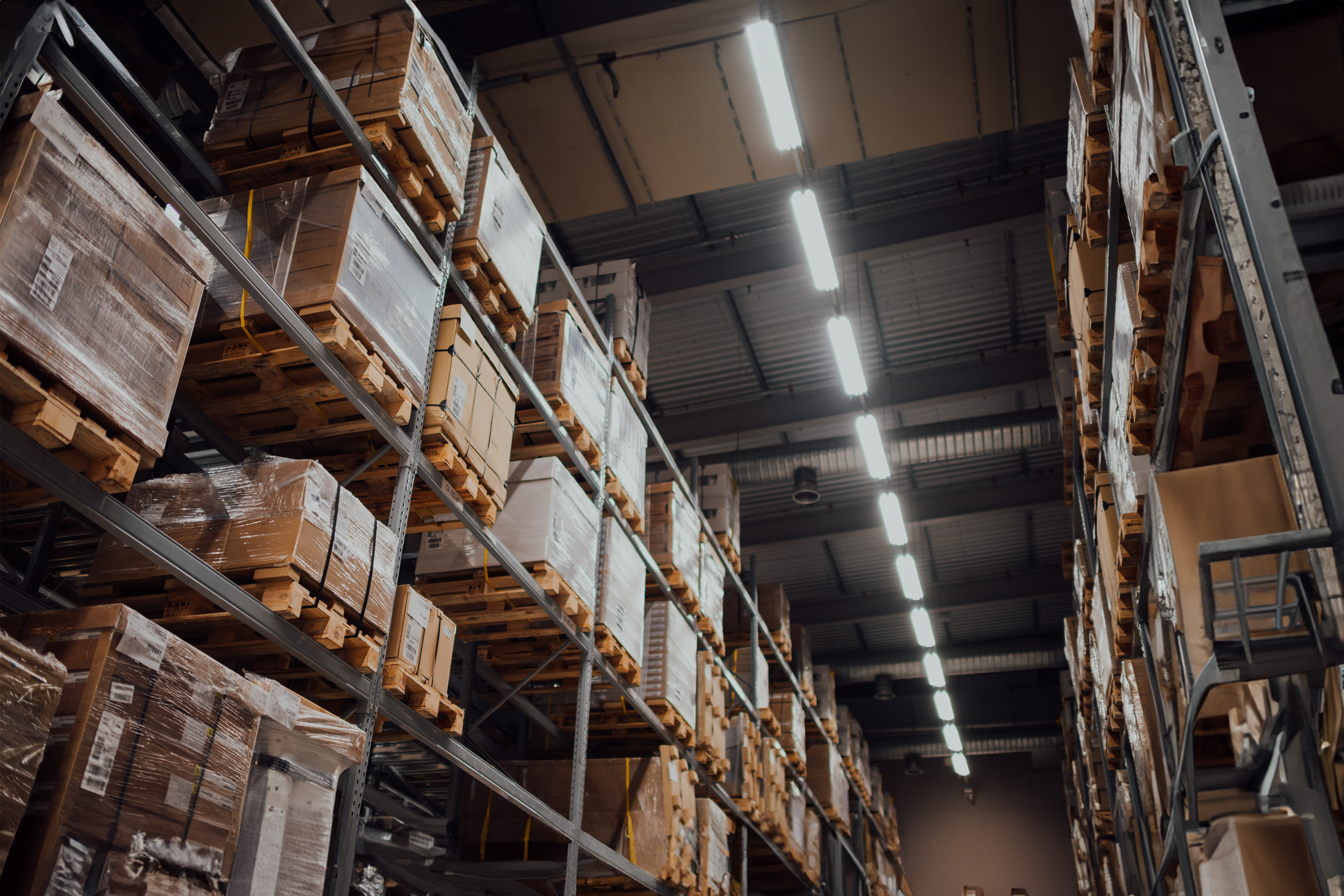 Warehouse interior with tall shelves stacked with boxed goods and illuminated by overhead lighting.