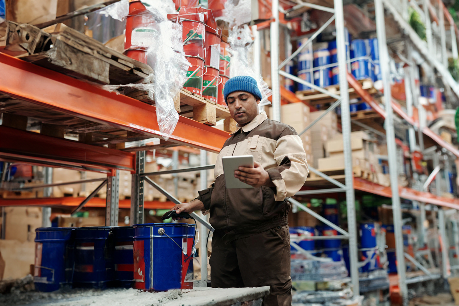 Worker in a warehouse using a tablet to manage inventory, surrounded by shelves stocked with various products.