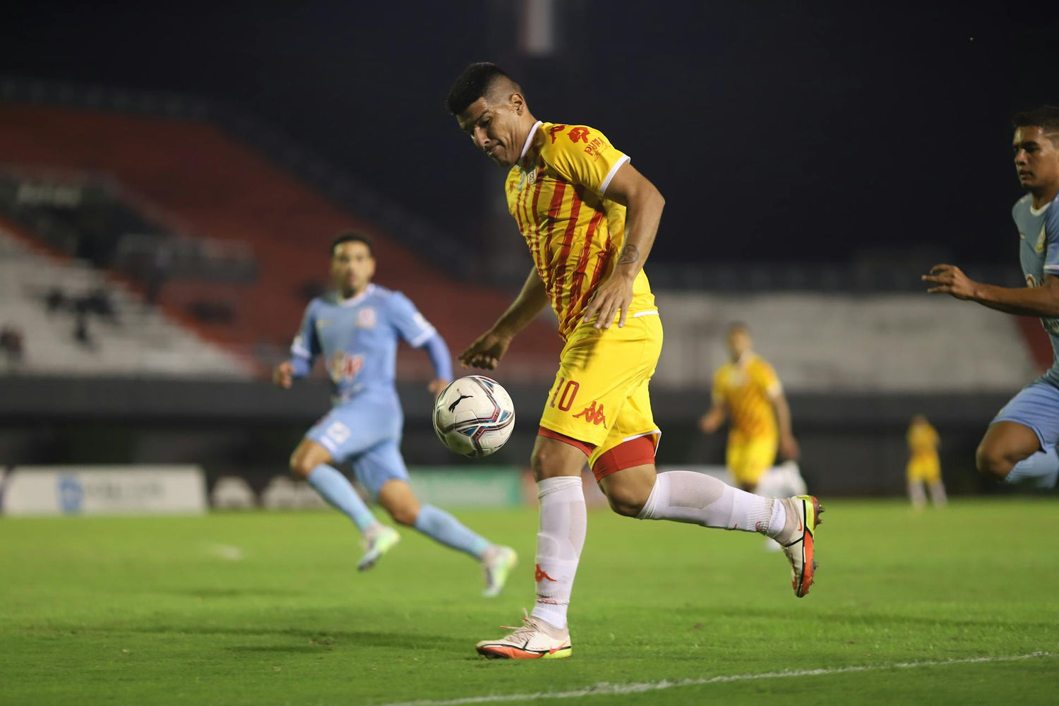 Soccer player in a yellow and red uniform by Trustline Enterprises, controlling the ball during a night match.