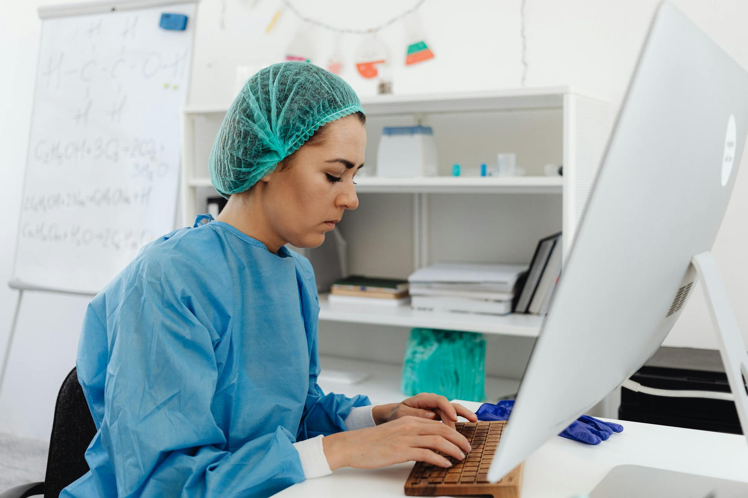 A healthcare professional in a blue gown and hairnet working on a computer in a clean and organized office environment, focusing intently on the task at hand.