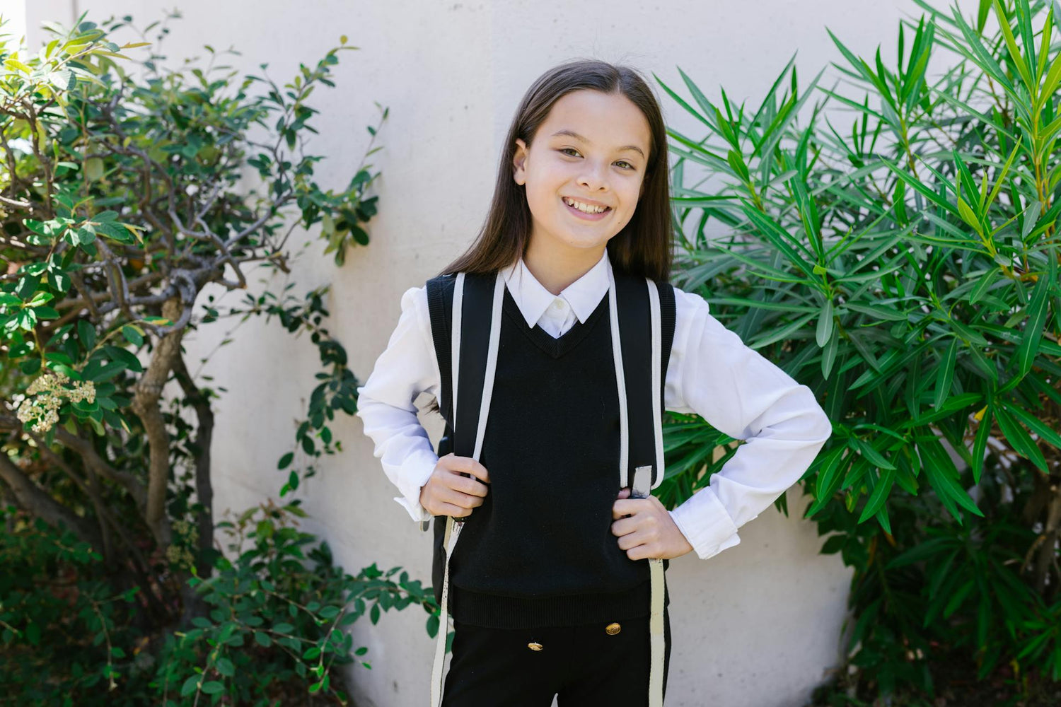 Smiling girl in a school uniform, wearing a black vest over a white shirt, holding her backpack straps, standing in front of a white wall with green foliage in the background.