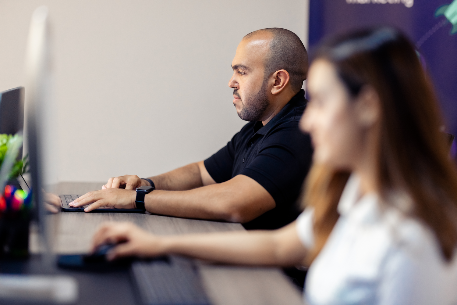 A person in a black shirt working intently on a computer, with a blurred background of a colleague also working on a computer.