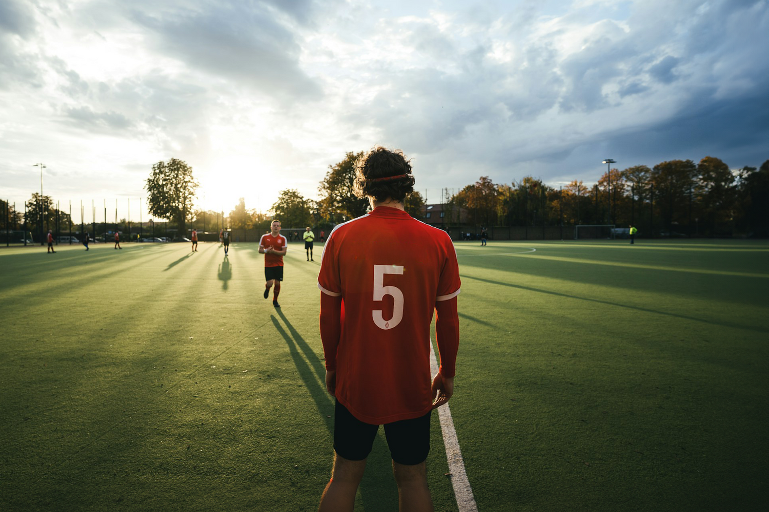 Soccer player wearing a red jersey with the number 5, standing on a green field at sunset, with teammates and trees in the background.