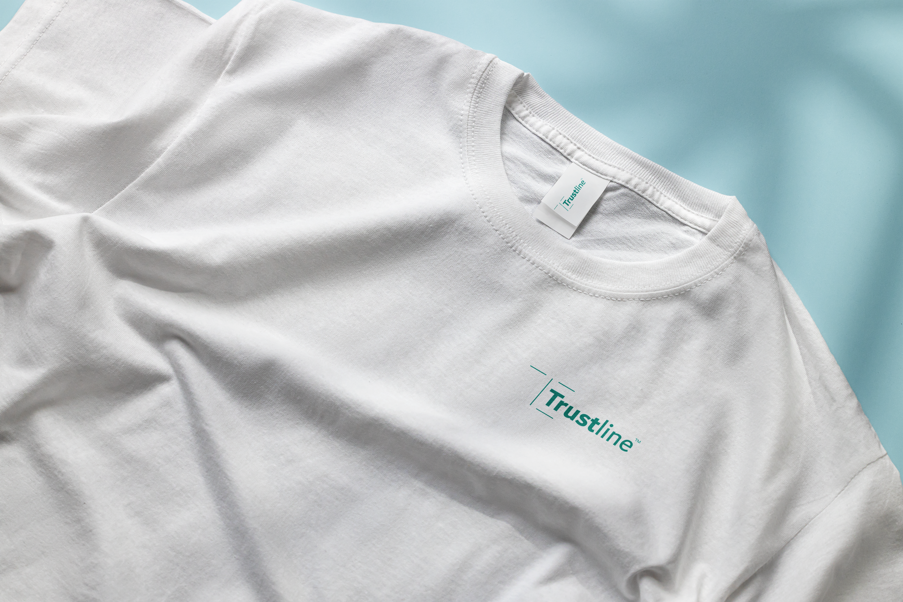 Close-up of a plain white t-shirt laid flat on a light blue background, featuring the Trustline logo on the chest and a neck tag with the Trustline branding.