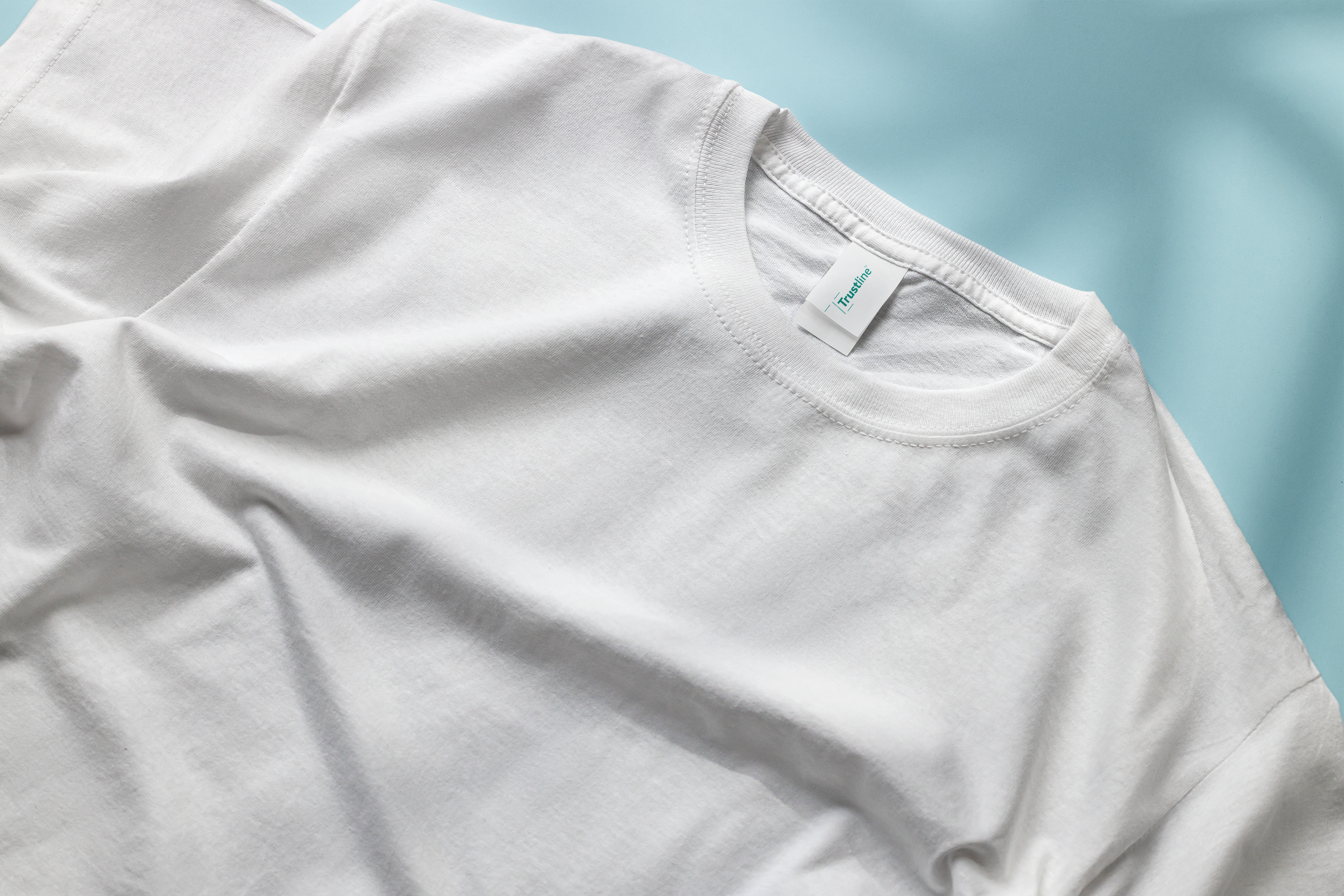 Close-up of a plain white t-shirt laid flat on a light blue background, with a visible neck tag featuring the Trustline logo.