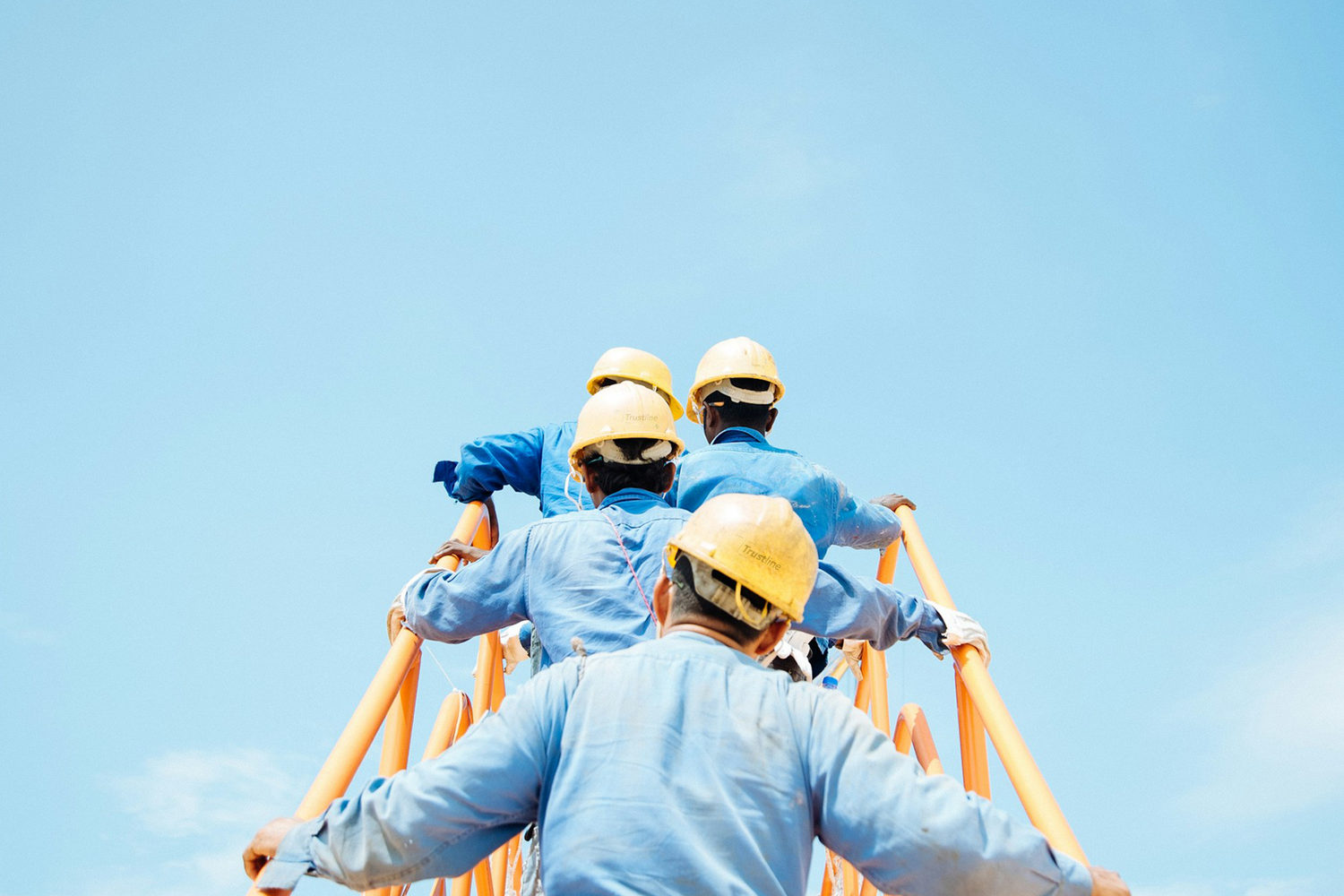 Construction workers in blue uniforms and yellow hard hats climbing an orange ladder against a clear blue sky, viewed from behind.